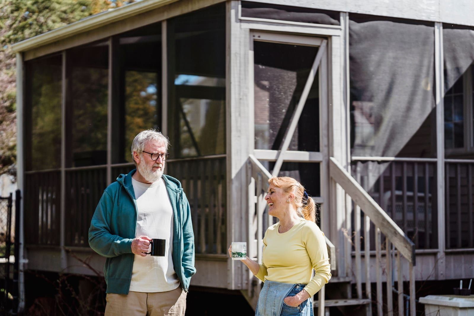 Content husband and wife in casual clothes standing with cups of tea near country house in sunlight.