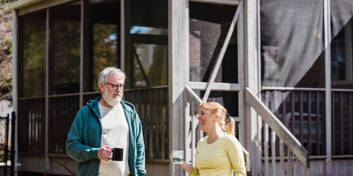 Content husband and wife in casual clothes standing with cups of tea near country house in sunlight.