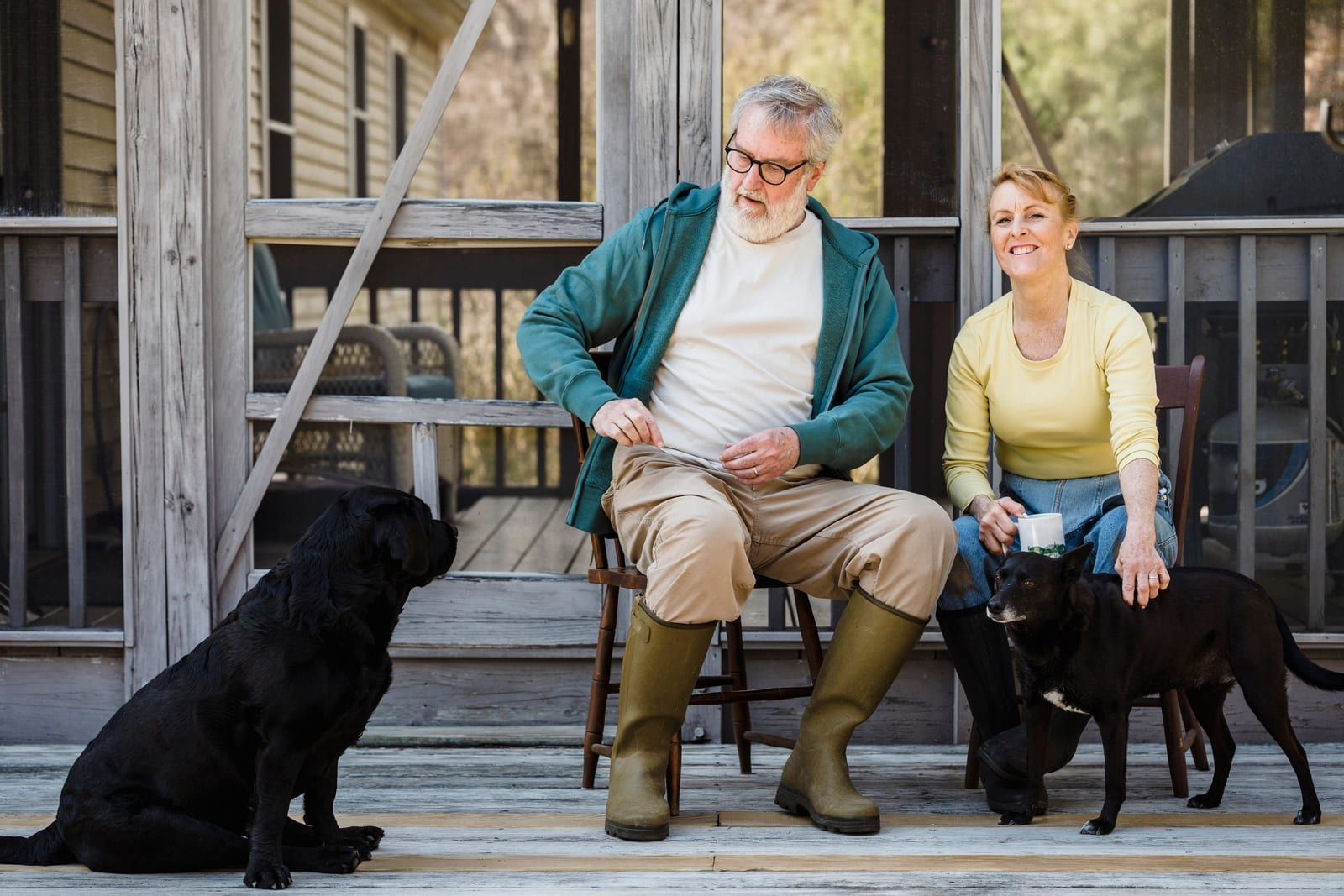 Delightful senior couple in rain boots sitting on chairs with black dogs while spending time together on terrace.