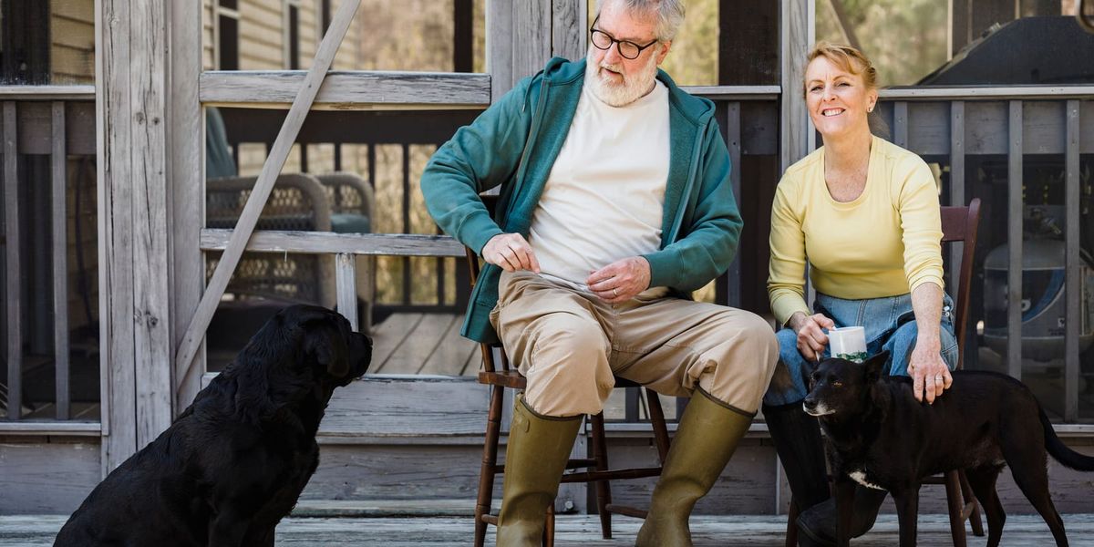 Delightful senior couple in rain boots sitting on chairs with black dogs while spending time together on terrace.